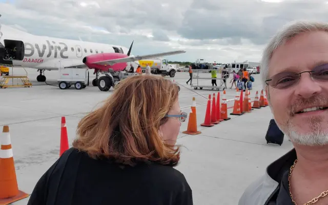 John and Lori selfie on airport tarmac with plane in background; both fully clothed