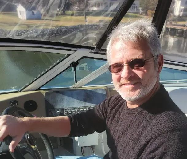 Man (John) smiling at boat helm wearing sunglasses and sweater; fully clothed