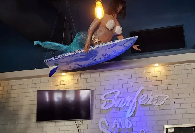 Decorative mermaid statue/prop with shell pasties surfing above bar; artwork/decor not a real person
