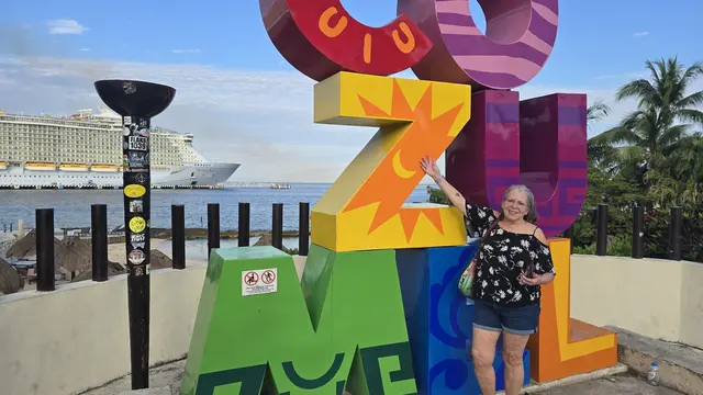 Lori posing at the colorful COZUMEL sign, cruise ship behind her.