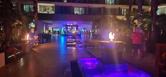 Resort courtyard glowing at night with fire bowls and neon palm trees.