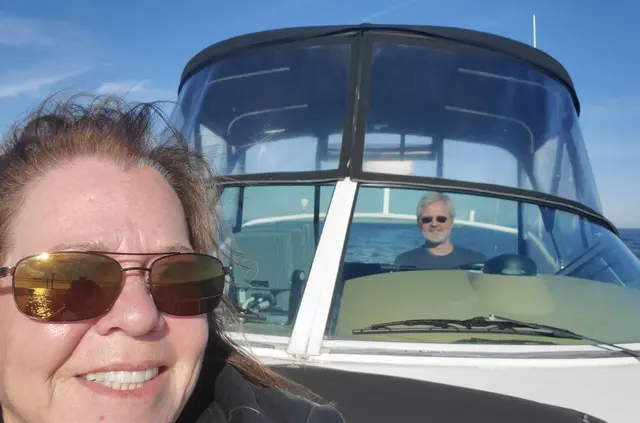 Woman (Lori) selfie with man (John) visible driving boat behind her; both fully clothed